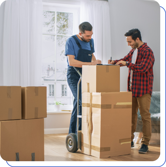 Two men in a room with large windows are handling cardboard boxes. One man, wearing overalls, stands by a hand truck, while the other signs a document. Several taped boxes are present.